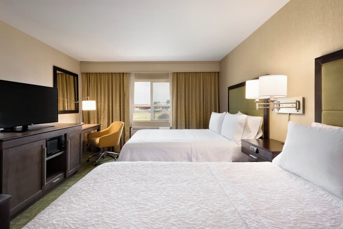 A hotel room with two beds, a desk with a flat-screen TV, a chair, and a window with curtains.