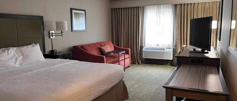 A hotel room with a large bed, a red armchair, a desk with a television, and an air conditioning unit.