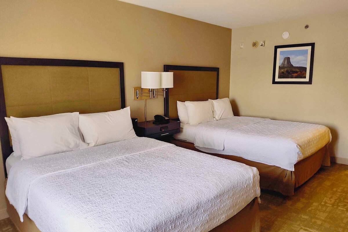 A hotel room with two beds, each with white linens and headboards. There is a bedside table with a lamp and a clock on the wall.