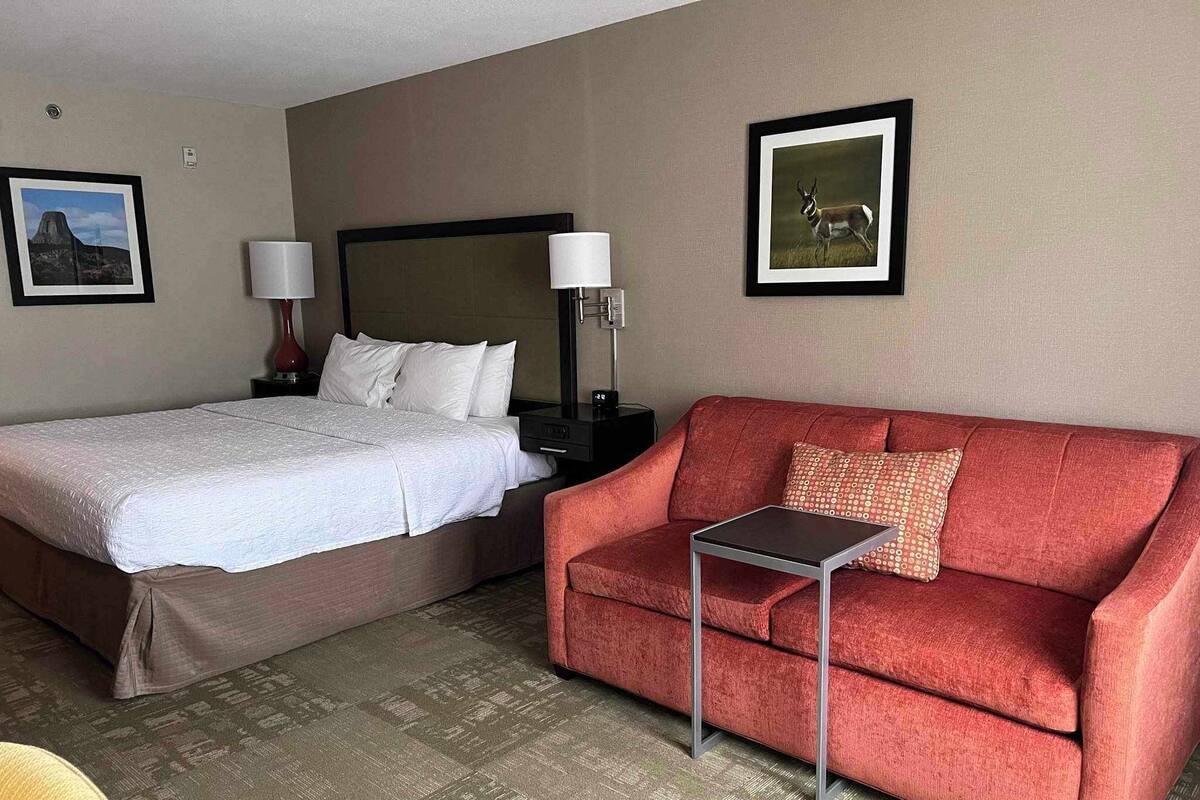 A hotel room with a bed, a red sofa, a small table, and two wall-mounted lamps.