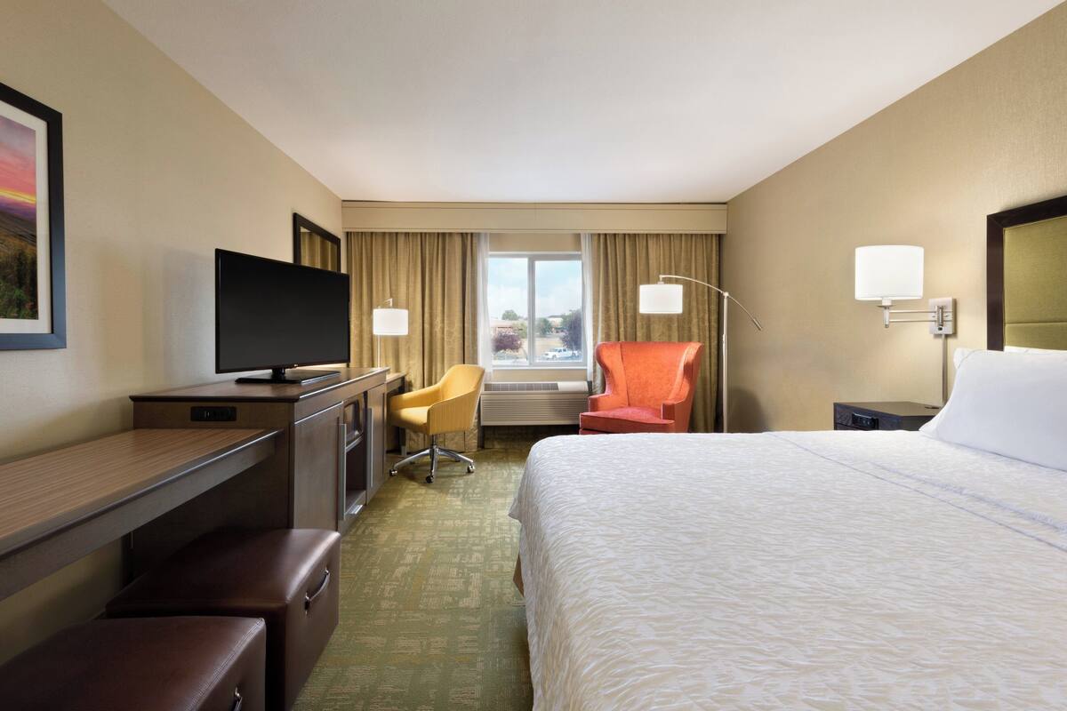 A hotel room with a bed, a desk with a television, a chair, a window with curtains, and a lamp.