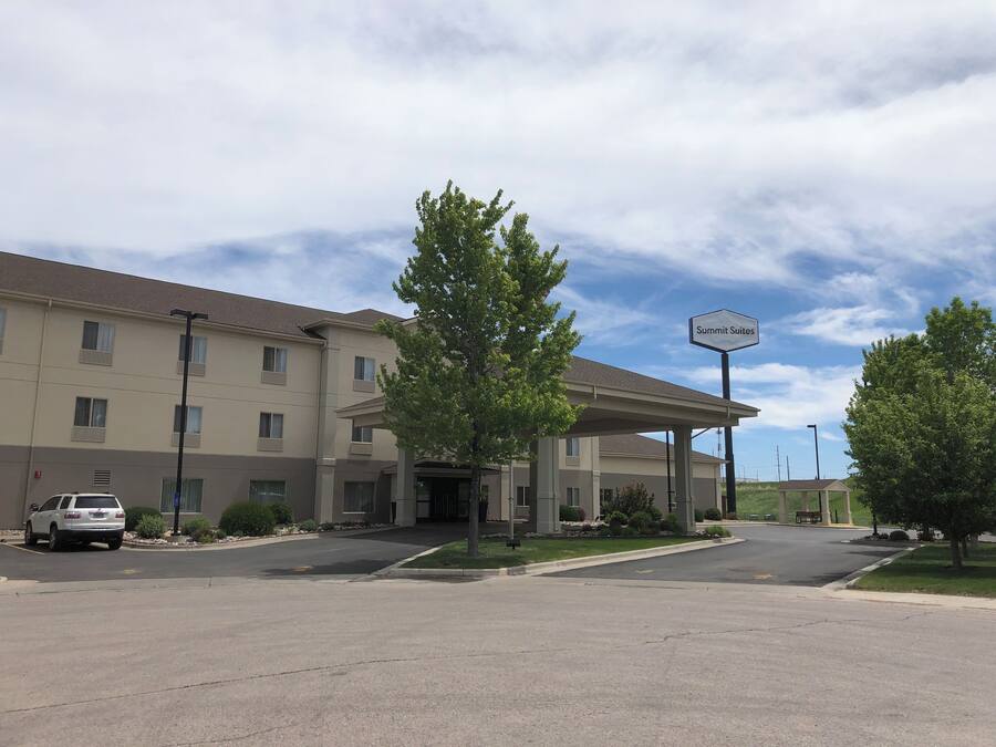 A hotel with a covered entrance, a sign indicating 'Summit Suites', and a parking lot with a white car.
