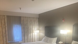 Una habitación de hotel con dos camas, una ventana con cortinas, un escritorio y una mesita de noche con lámpara.