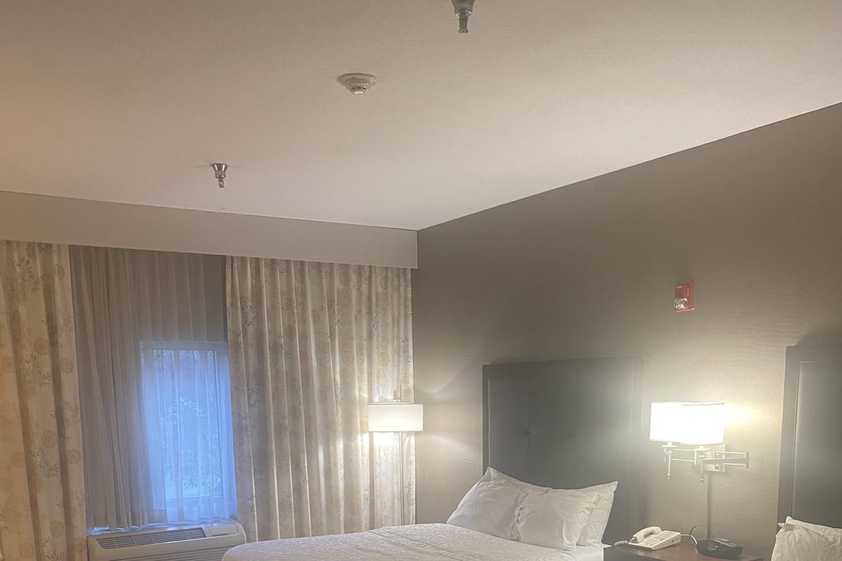 Una habitación de hotel con dos camas, una ventana con cortinas, un escritorio y una mesita de noche con lámpara.
