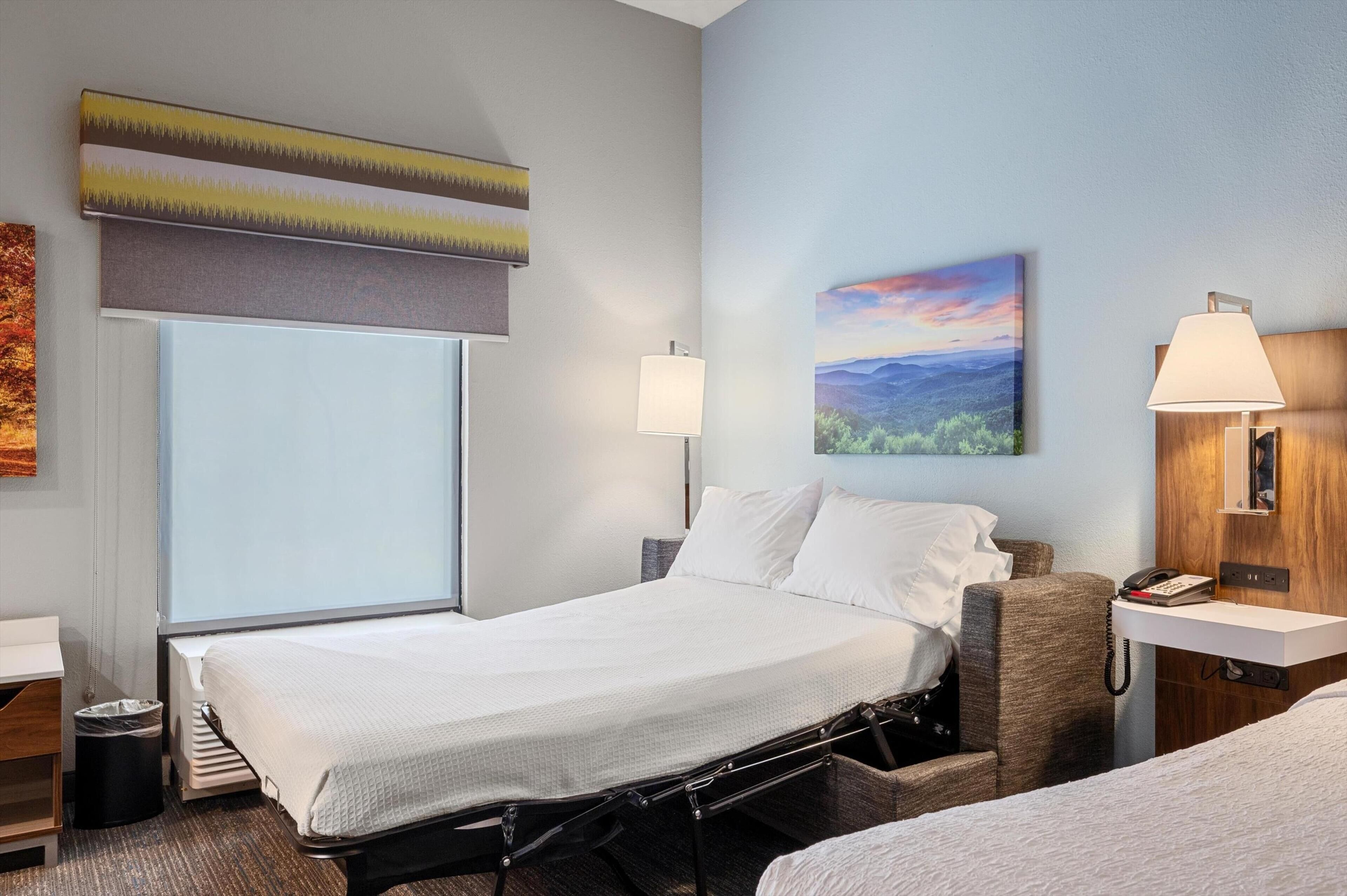 Een hotelkamer met een bed, nachtlampjes, een schilderij en een raam met jaloezieën.