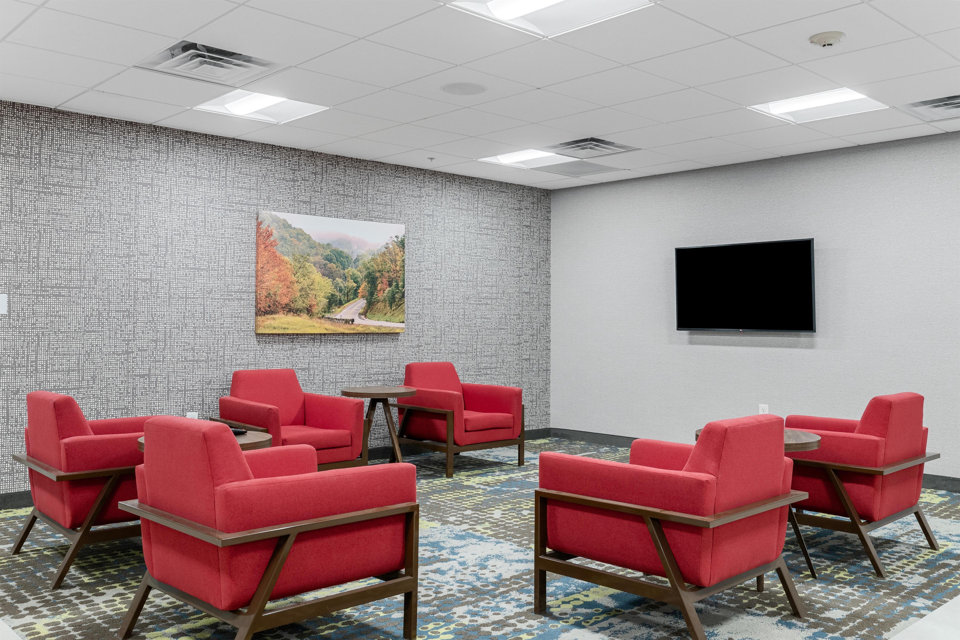 A conference room with red chairs, a round table, a painting of a landscape, a flat-screen TV, and patterned carpet.