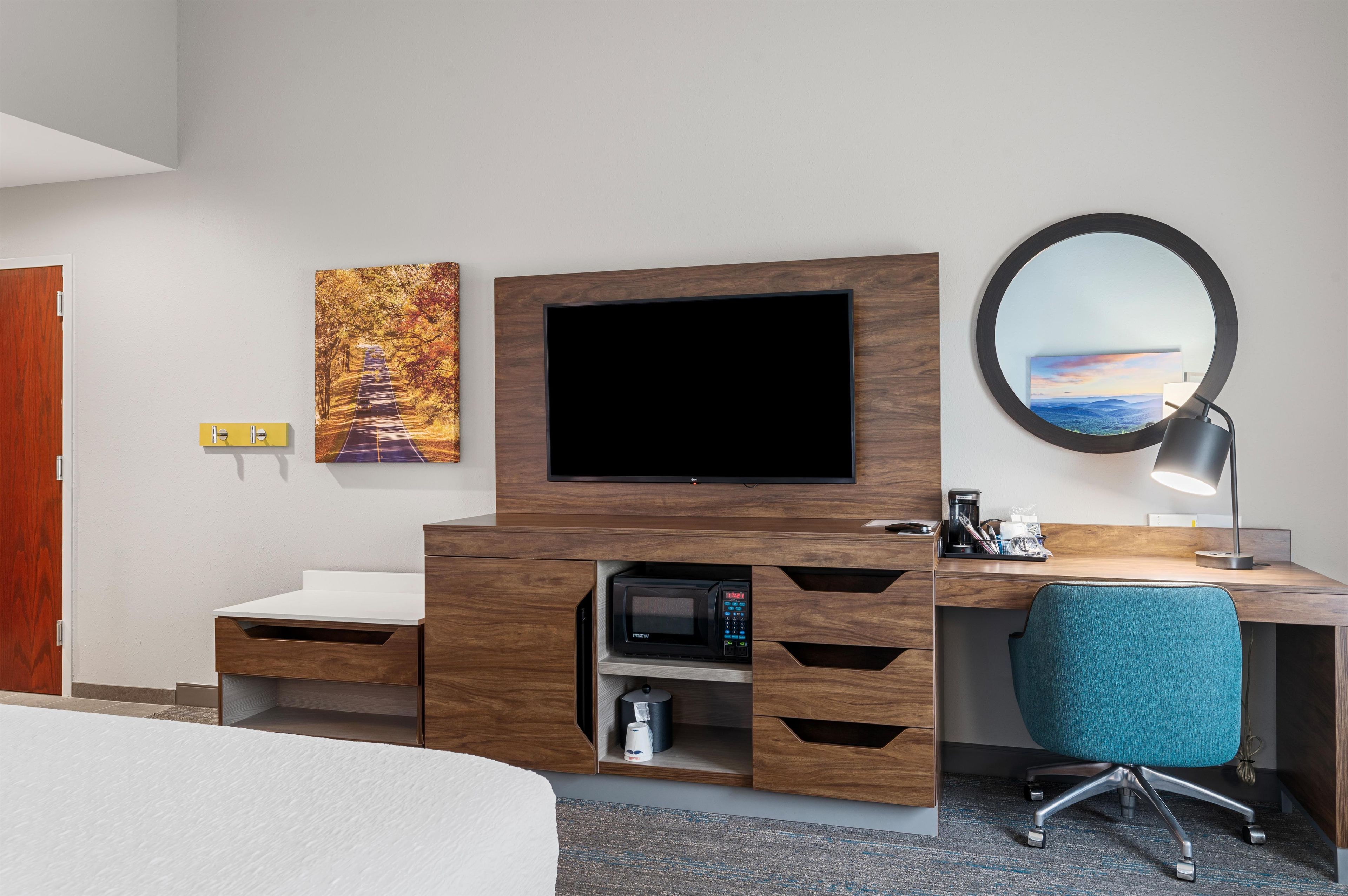 A hotel room with a flat-screen TV, a desk with a coffee maker, a chair, and a painting on the wall.