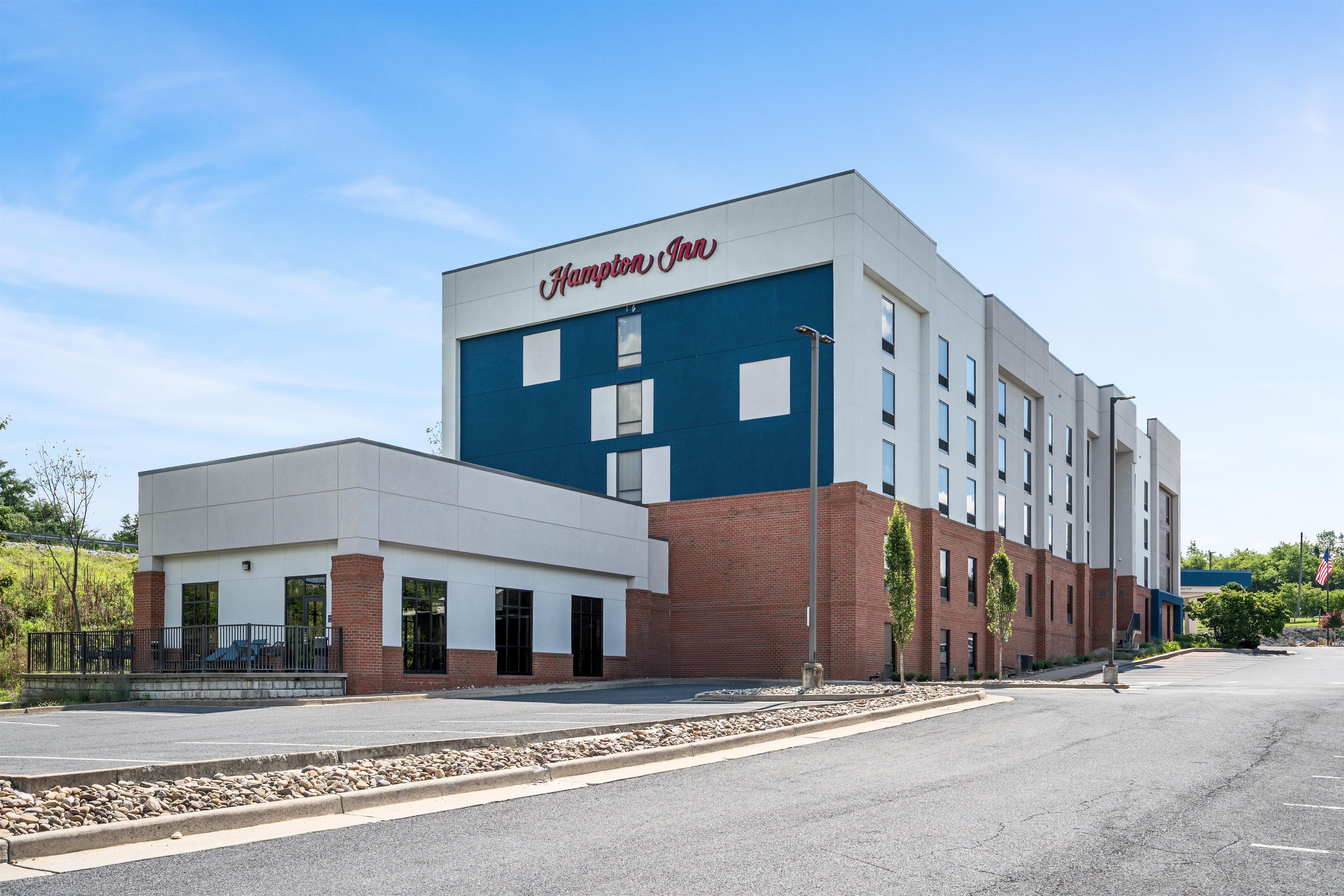 Un hotel Hampton Inn con diseño moderno, que presenta una fachada destacada en azul y blanco, un estacionamiento y un asta de bandera.