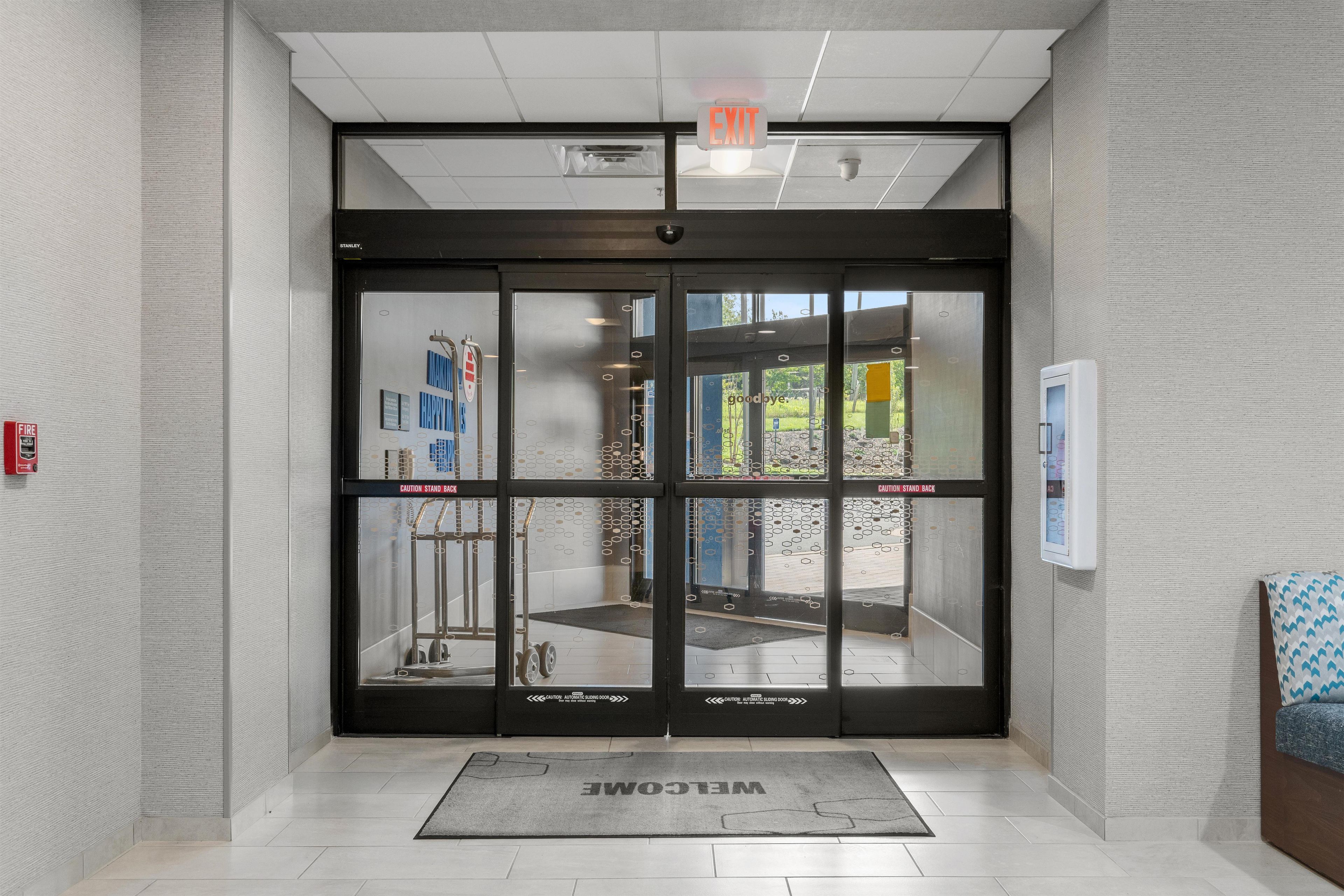 A modern entrance with a glass door, a welcome mat, and a fire alarm on the wall.
