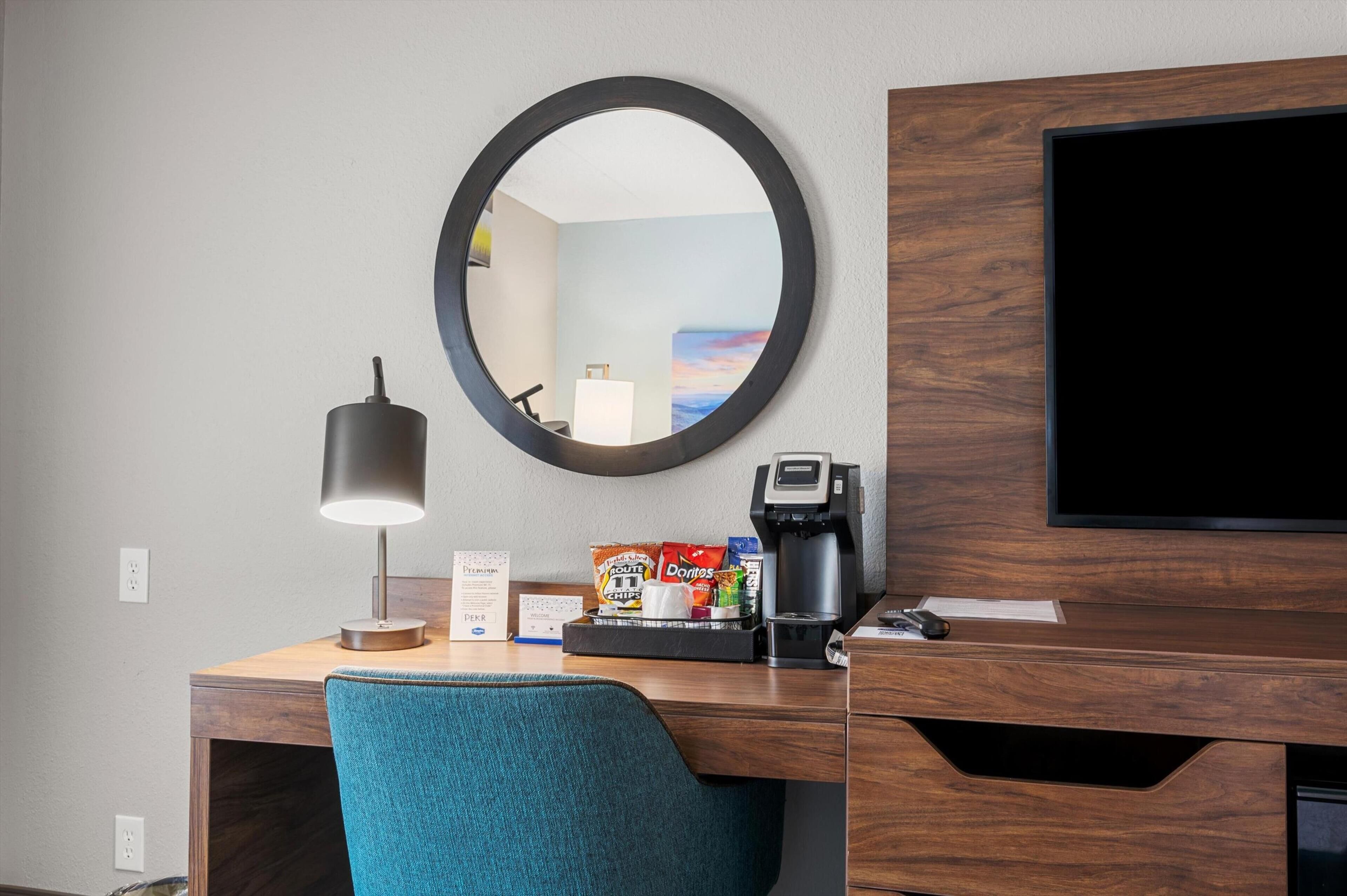 A hotel desk with a round mirror, a lamp, a coffee maker, snacks, and a TV.