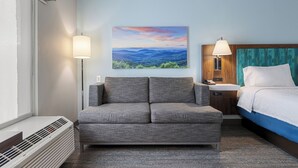 A hotel room with a gray sofa, a bed with a blue headboard, a painting of a landscape, and a lamp on the nightstand. - Hampton Inn Front Royal (Front Royal)