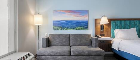 A hotel room with a gray sofa, a bed with a blue headboard, a painting of a landscape, and a lamp on the nightstand.