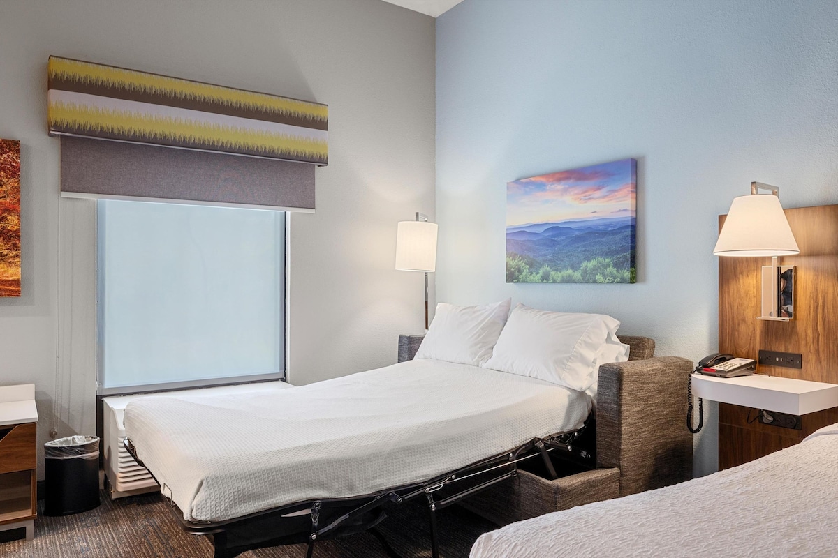 A hotel room with a bed, bedside lamps, a painting, and a window with blinds.