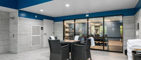 A modern indoor space with a dining area, a clock, and a view into a gym area.