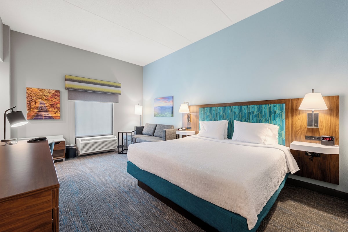 Ein Hotelzimmer mit einem großen Bett, einem Schreibtisch, einem Stuhl, einem kleinen Tisch und einem Nachttisch mit Lampe.