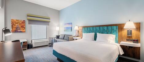 A hotel room with a large bed, a desk, a chair, a small table, and a nightstand with a lamp.