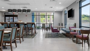 A modern dining area with tables and chairs, large windows, and a flat-screen TV on the wall. - Hampton Inn Front Royal (Front Royal)
