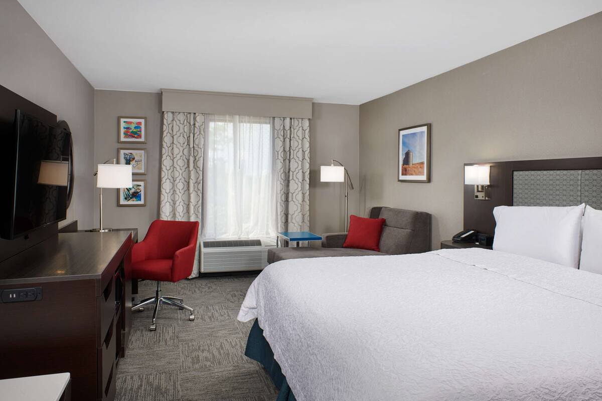 A hotel room with a large bed, a desk with a flat-screen TV, a red armchair, a gray armchair, a small table, and a window with patterned curtains.