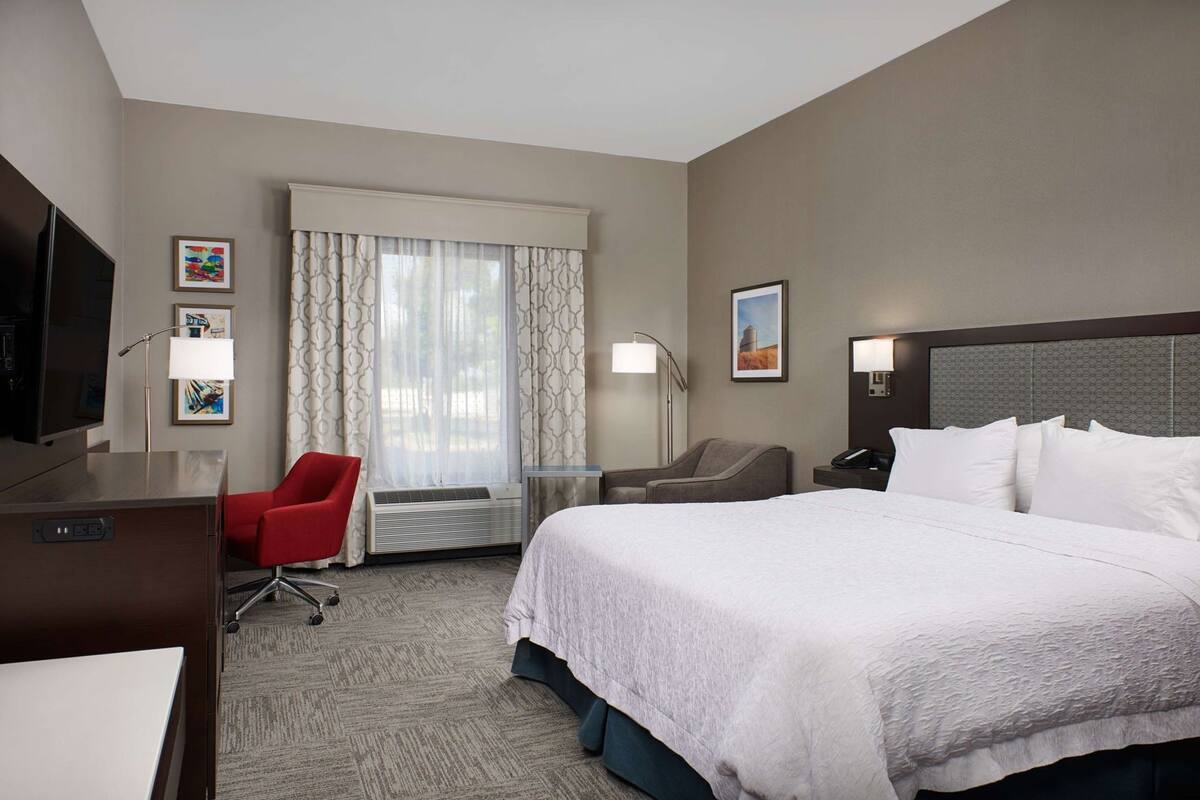 A hotel room with a bed, a desk, a chair, a TV, and a window with curtains.