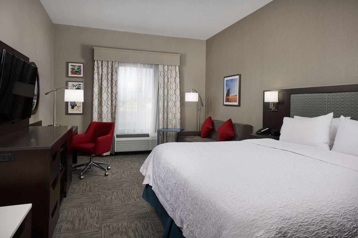 A hotel room with a large bed, a desk with a chair, a TV, a window with curtains, and a wall with framed pictures.