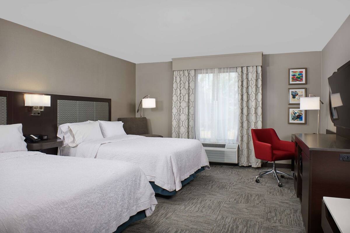 A hotel room with two beds, a desk, a red chair, and a large window with curtains.
