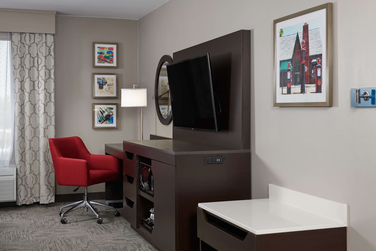 A hotel room with a desk, a red office chair, a flat-screen TV, a mirror, and framed artwork on the wall.