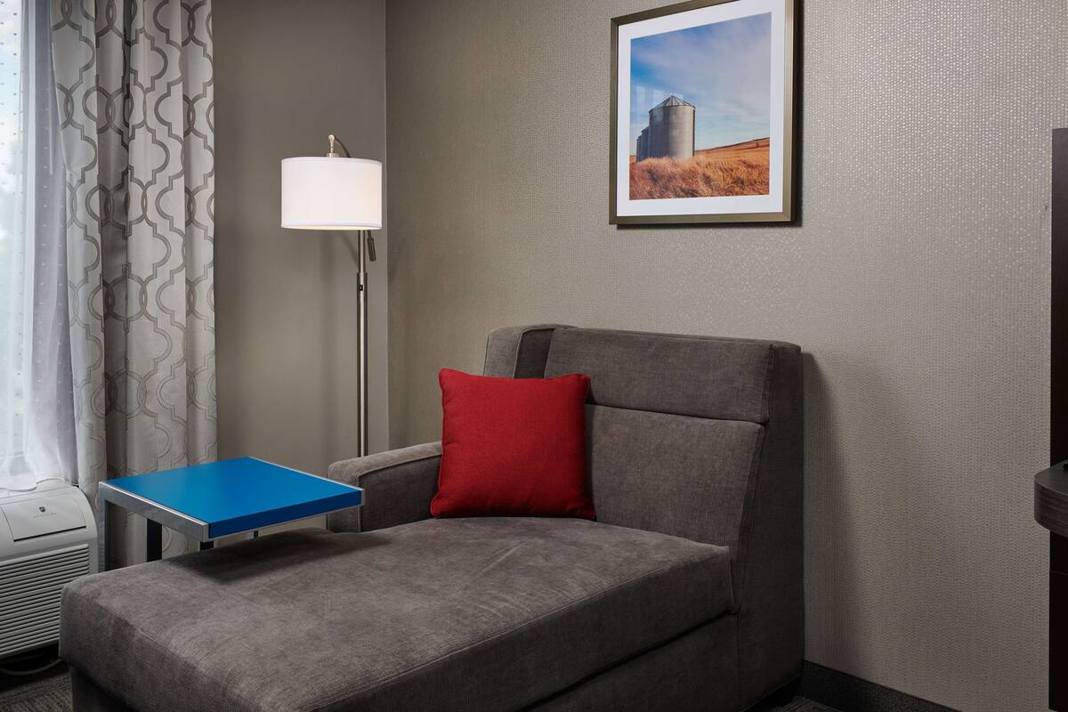 A hotel room with a grey sofa, a blue table, a red pillow, a floor lamp, and a framed picture on the wall.