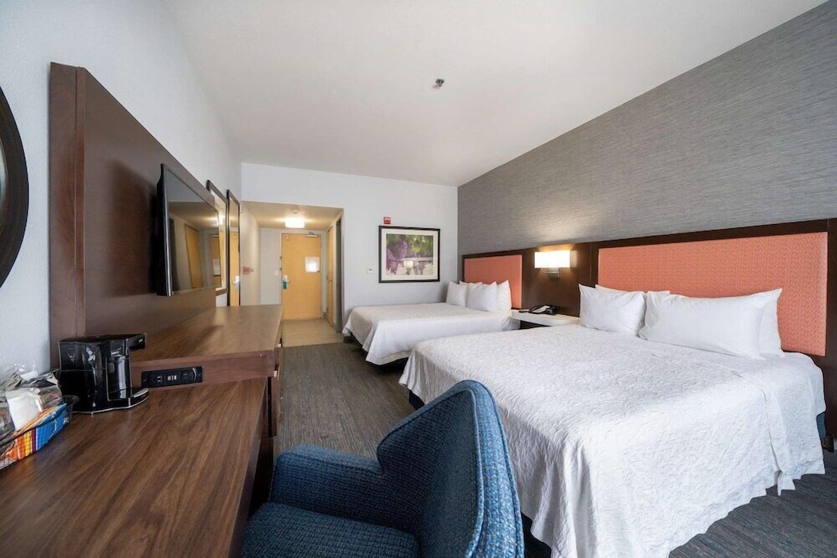 A hotel room with two beds, a desk with a coffee maker, a chair, a large mirror, and a framed picture on the wall.