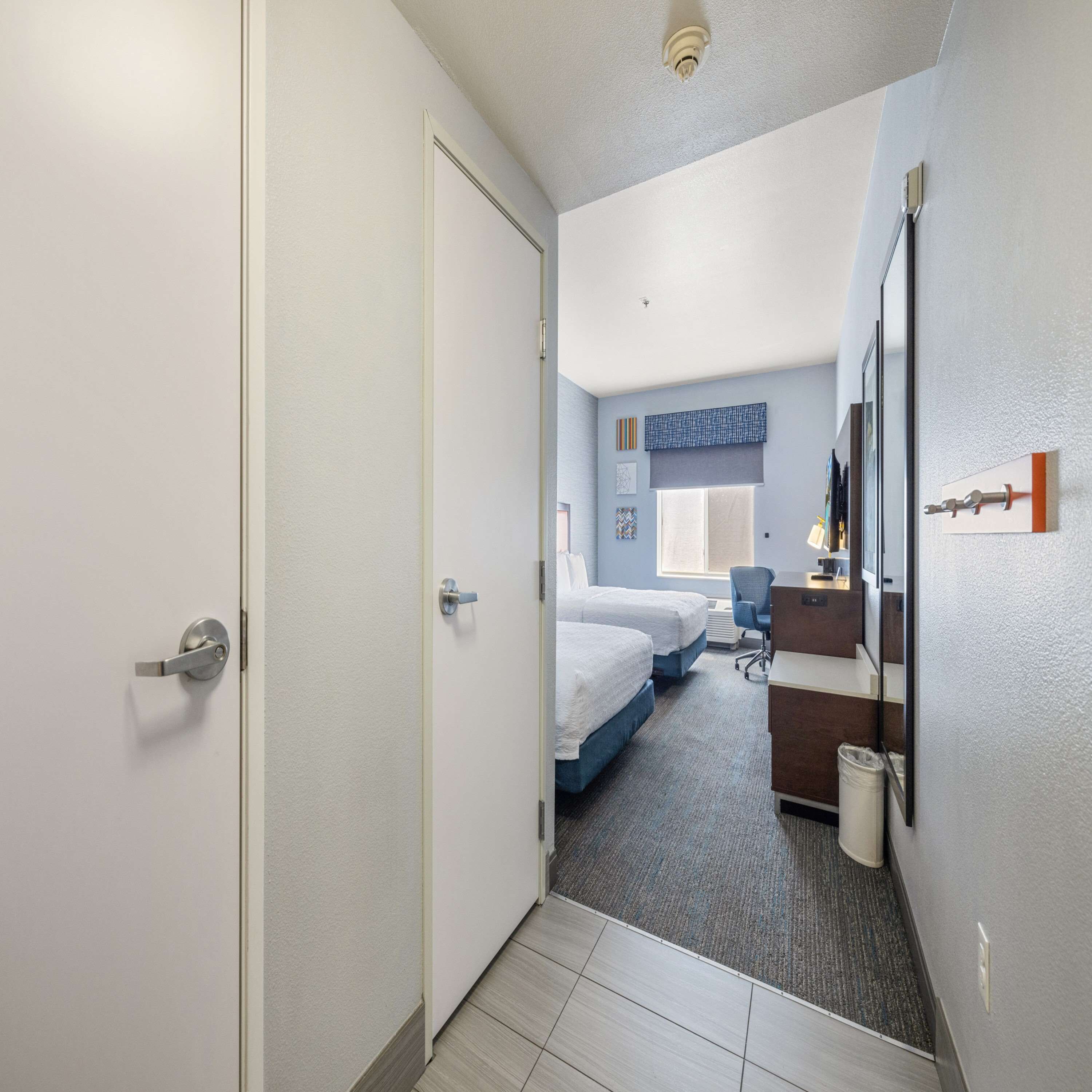 Habitación de hotel con puerta, dos camas, escritorio, silla, cubo de basura y ventana con persianas.