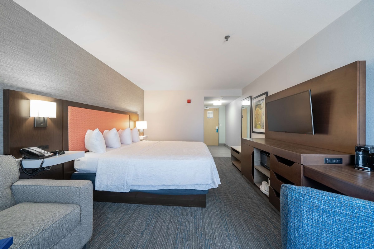 A hotel room with a bed, bedside table, lamp, TV, and a desk with a chair.