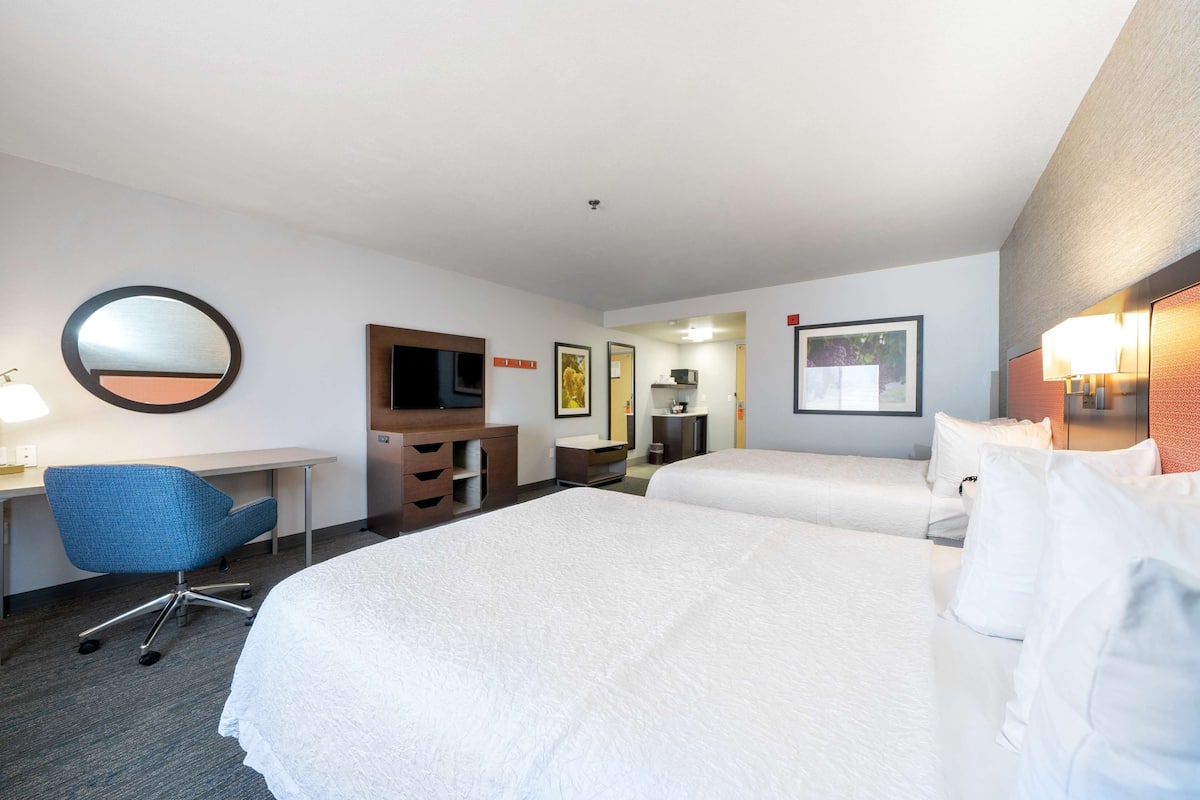 A hotel room with two beds, a desk with a chair, a TV, and a bathroom visible through an open door.