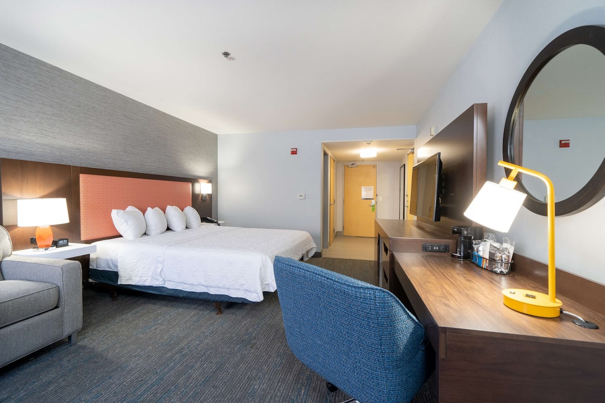 A hotel room with a bed, a desk with a lamp, a chair, a television, and a mirror.