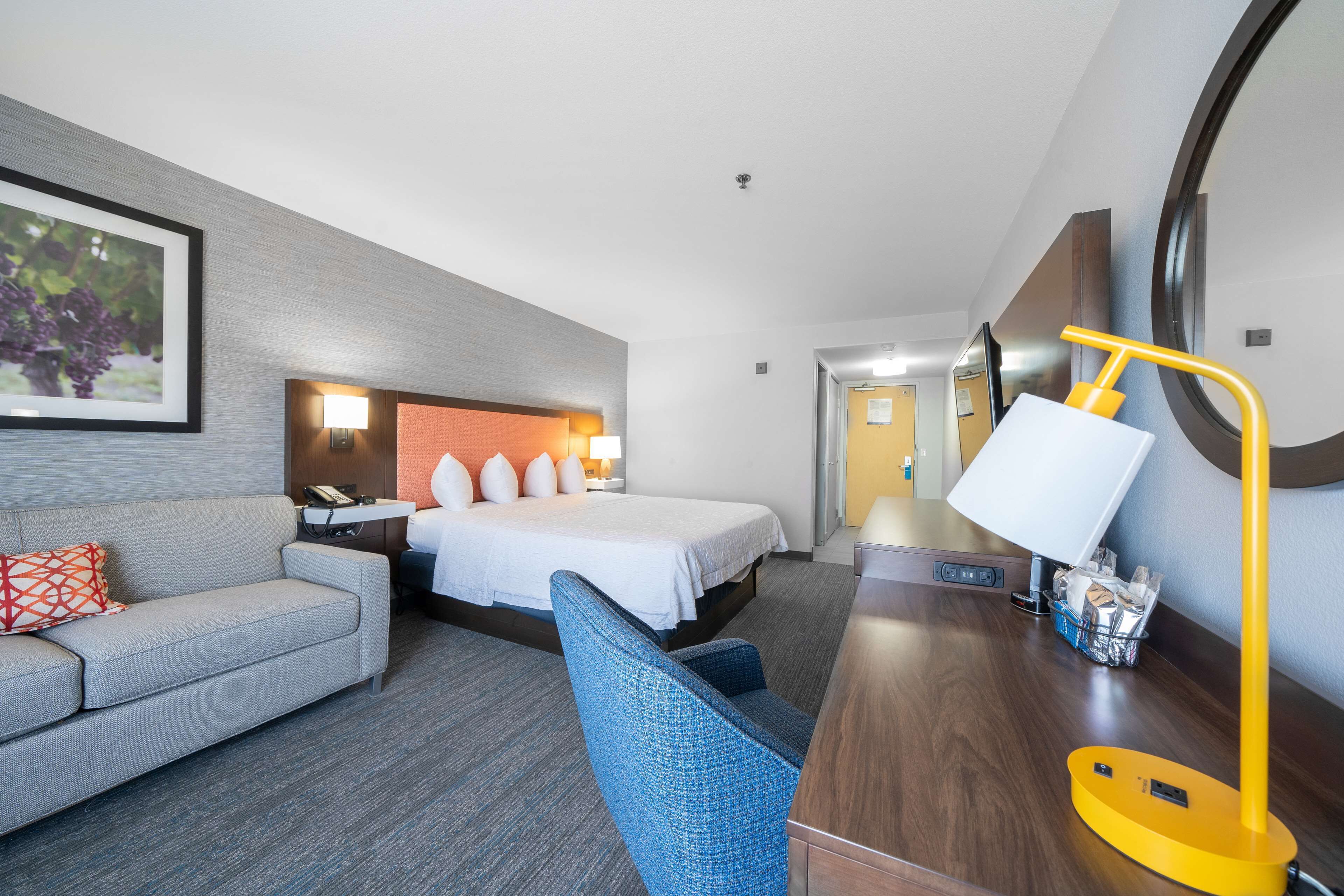 Habitación de hotel con cama, escritorio con lámpara, sillón, sofá y televisor montado en la pared.