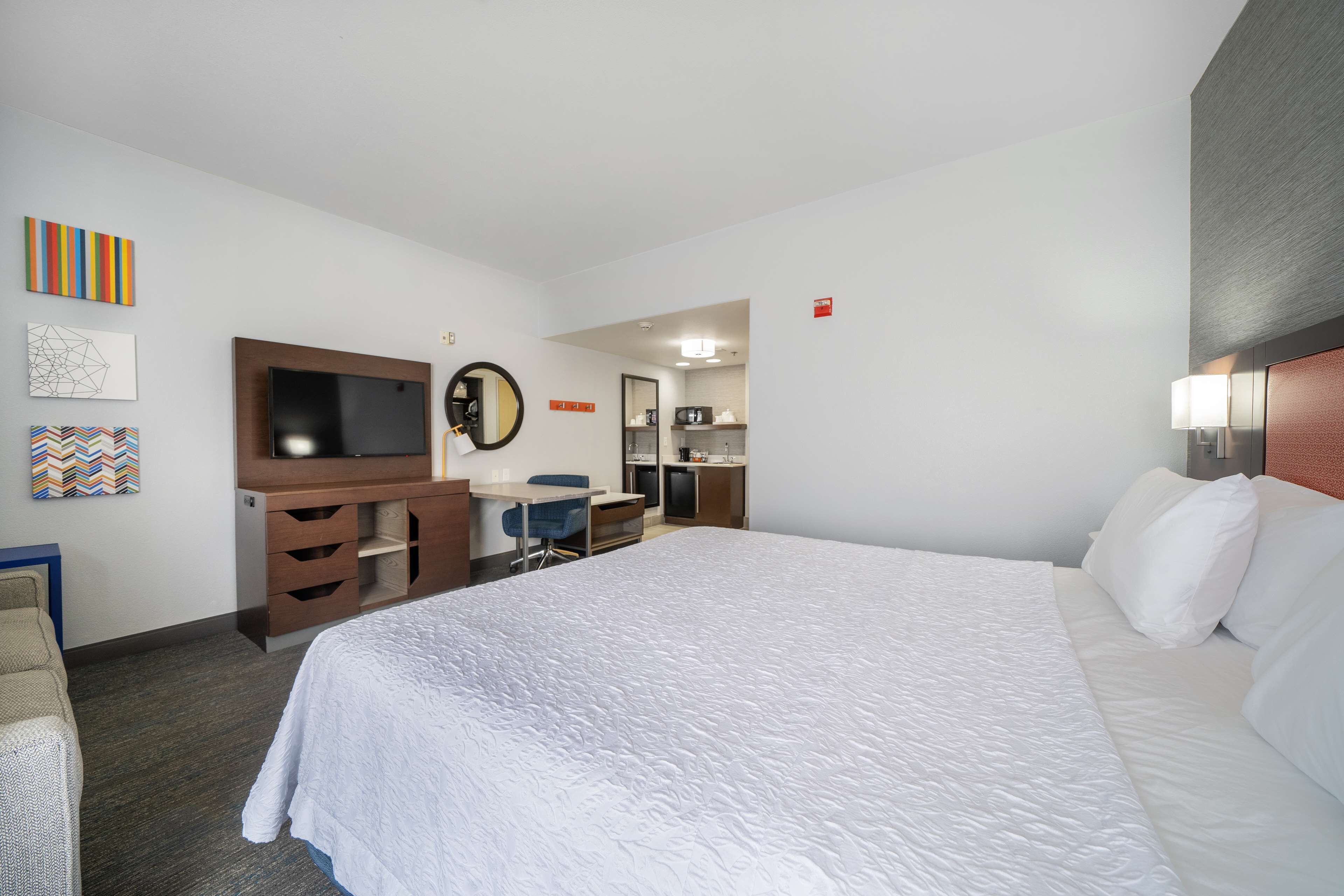 A hotel room with a large bed, a desk with a chair, a television, and a kitchenette in the background.