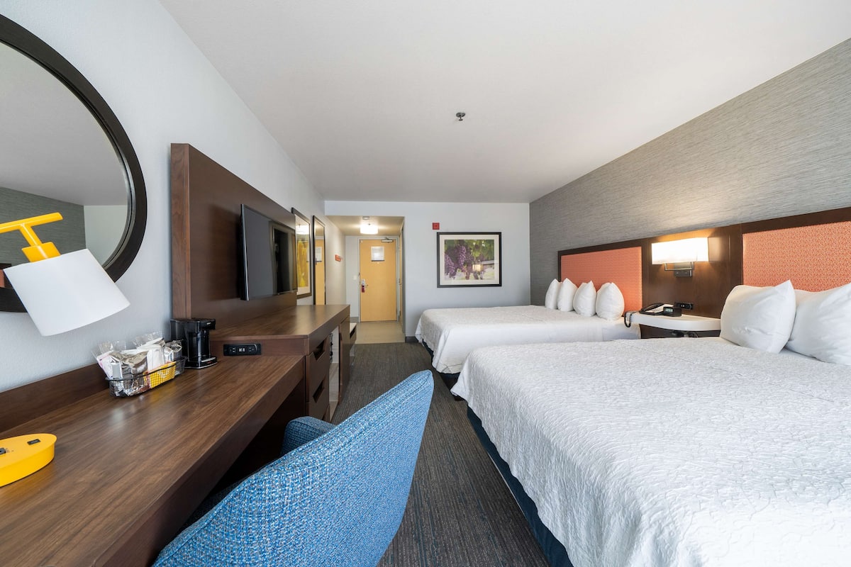 A hotel room with two beds, a desk with a chair, a TV, and a bathroom visible in the background.