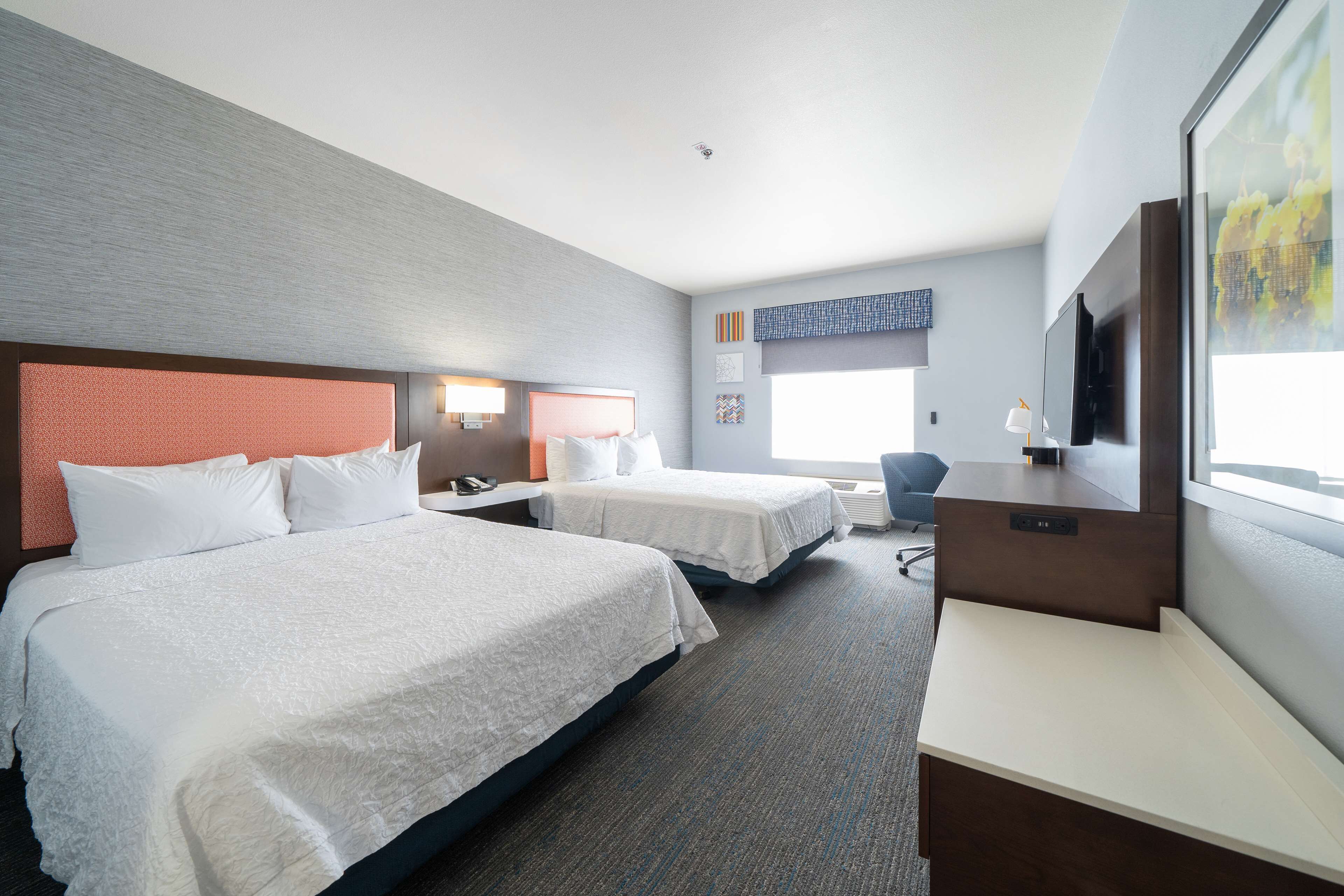 A hotel room with two beds, a desk, a chair, a TV, and artwork on the walls.
