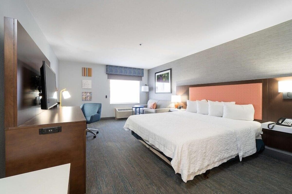 A hotel room with a large bed, a desk with a flat-screen TV, a chair, a desk lamp, and a window with blinds.
