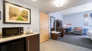 Quarto de hotel com cozinha compacta, cama, escrivaninha, cadeira, televisão e um quadro na parede.