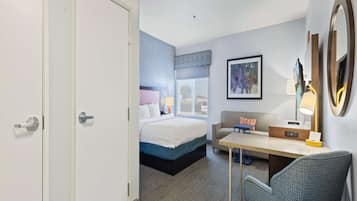 A hotel room with a bed, a desk, a chair, a sofa, a desk lamp, a mirror, and a framed picture on the wall.