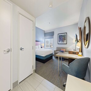 A hotel room with a bed, a desk, a chair, a sofa, a desk lamp, a mirror, and a framed picture on the wall.