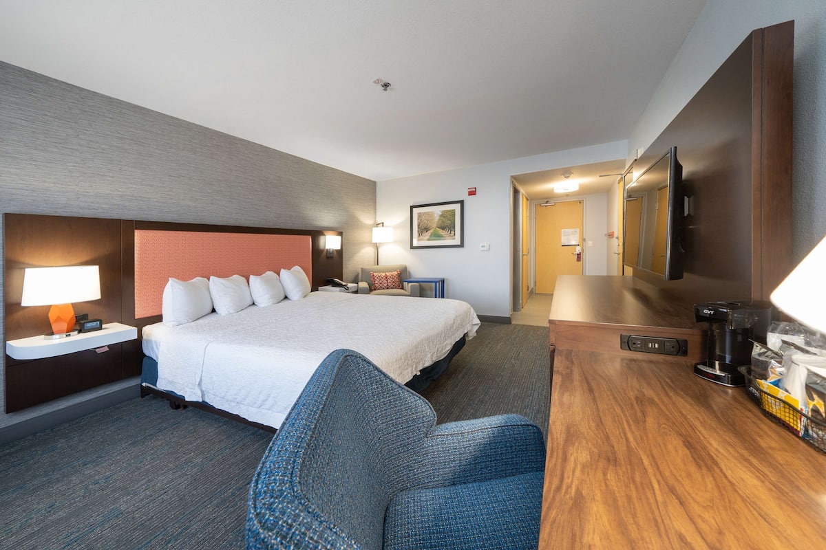 A hotel room with a bed, a desk with a coffee maker, a chair, a lamp, and a TV.