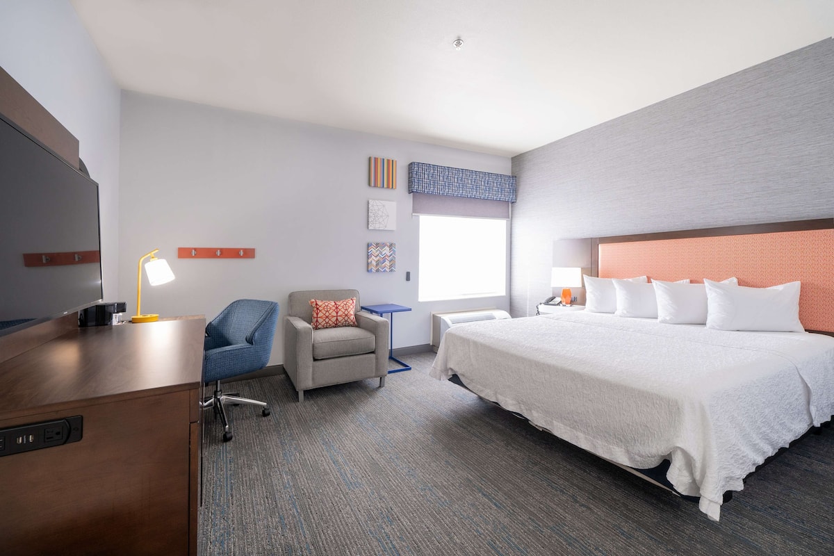 A hotel room with a large bed, a desk with a lamp, a chair, and a small table.
