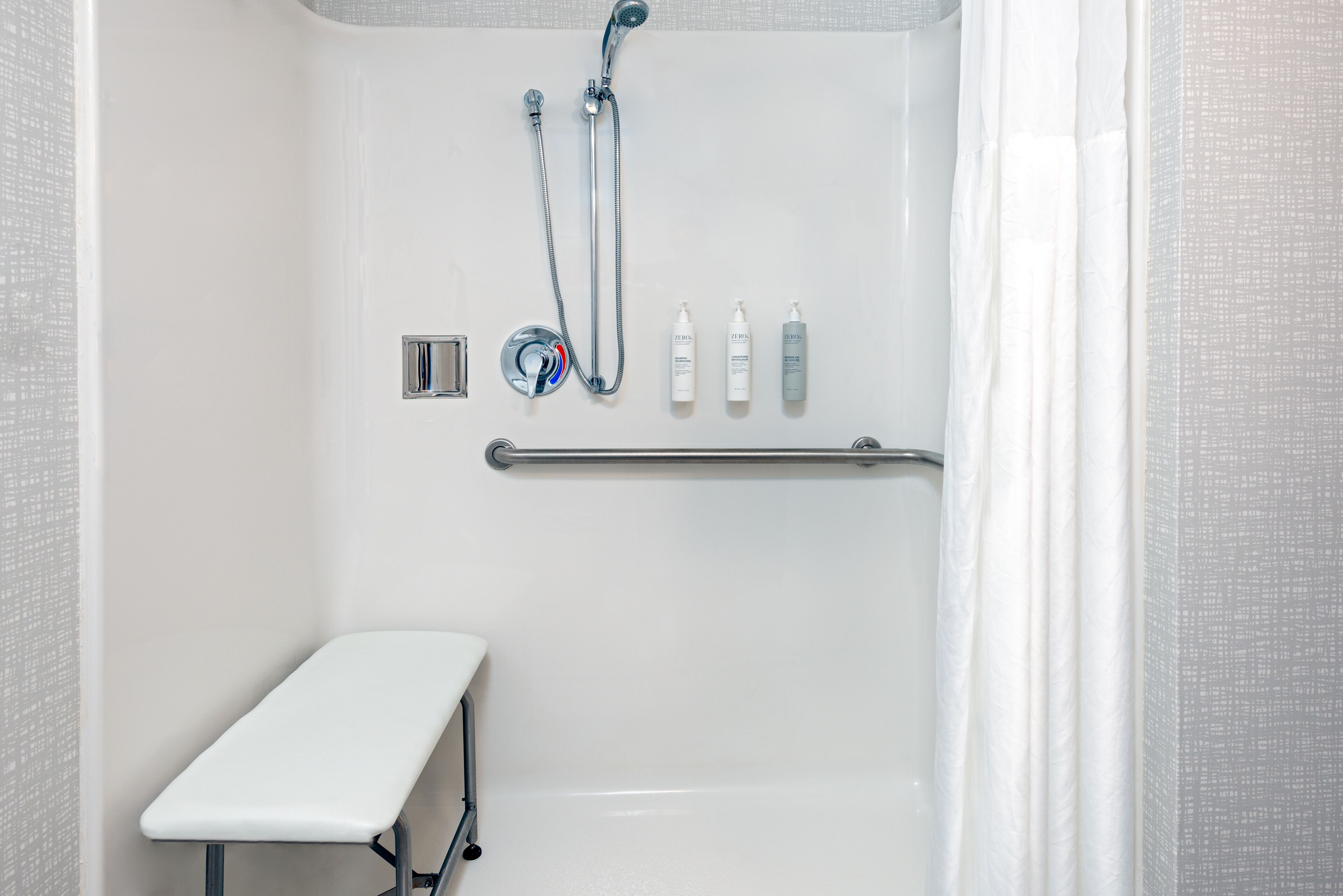 A white shower with a handheld showerhead, a handheld shower attachment, and a handheld shower nozzle. There is a white bench in the corner and a towel rack on the wall.