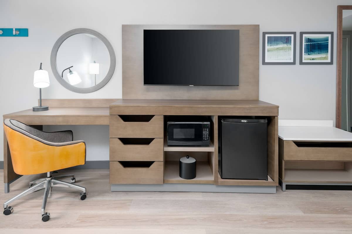 A modern hotel room with a wooden desk, a flat-screen TV, a microwave, a refrigerator, a chair, and a trash can.