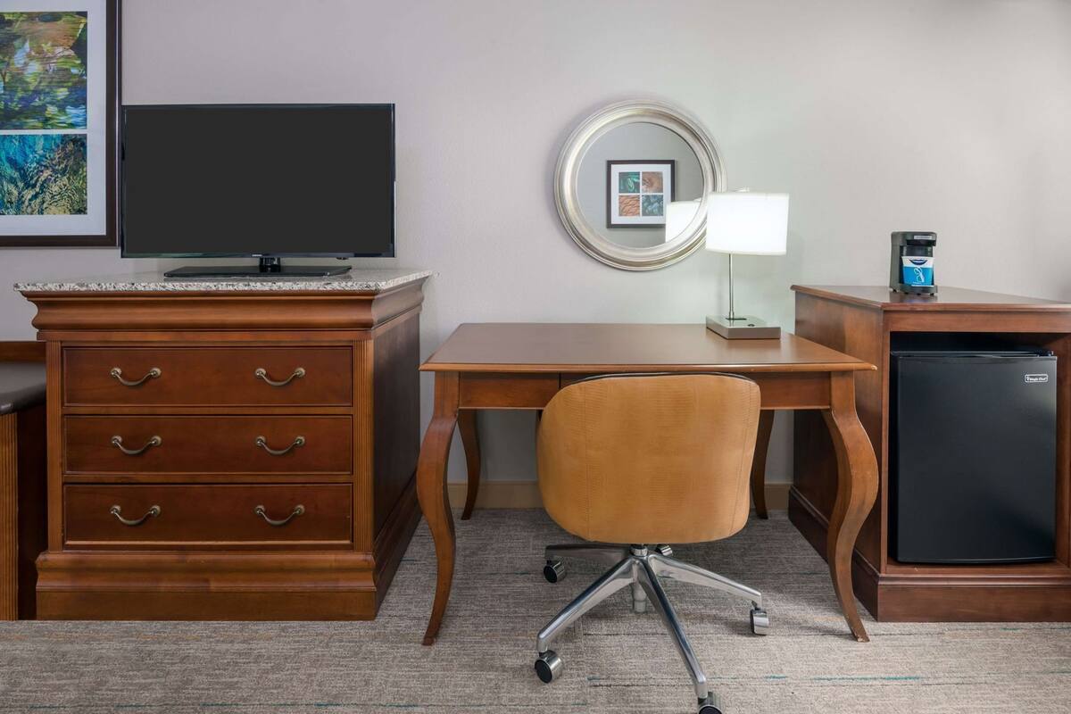 A hotel room with a wooden desk, a chair, a television, a refrigerator, a dresser, and a mirror.