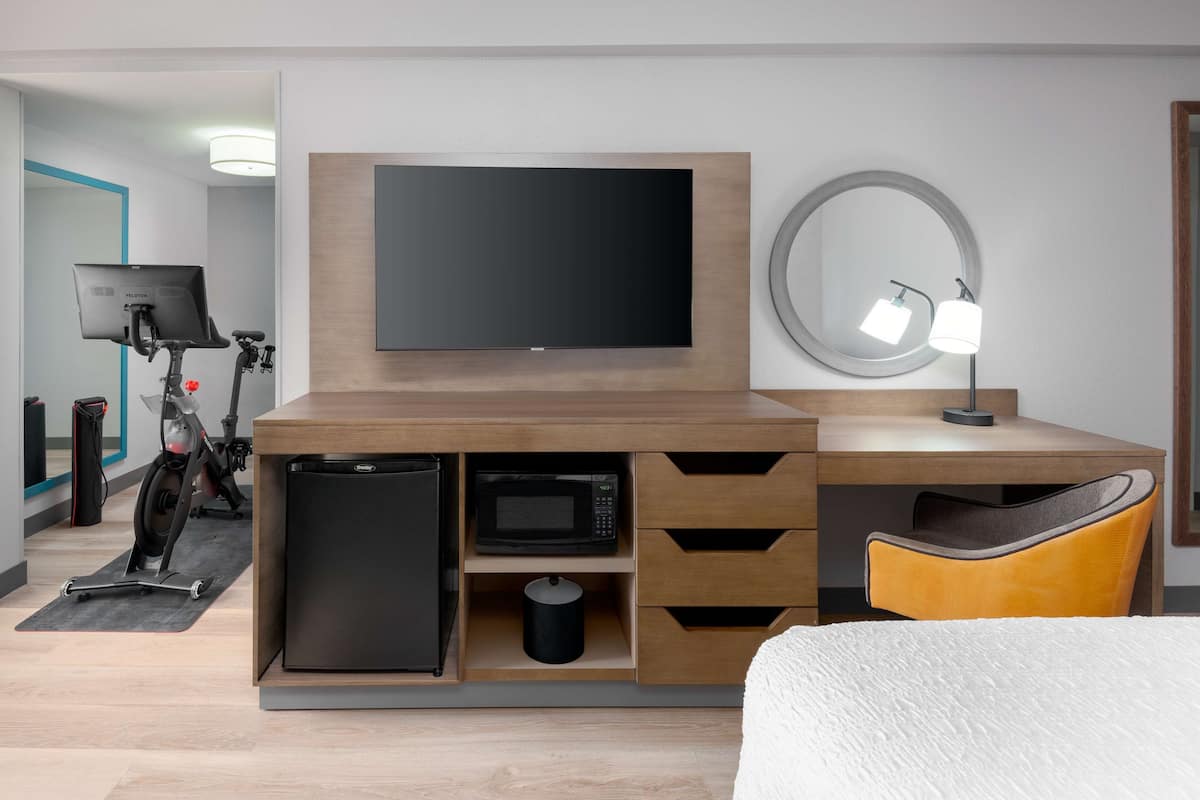 A modern hotel room with a wooden desk, a flat-screen TV, a microwave, a refrigerator, a small trash bin, a yellow chair, and a round mirror with a light.