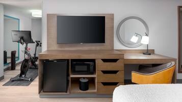 A modern hotel room with a wooden desk, a flat-screen TV, a microwave, a refrigerator, a small trash bin, a yellow chair, and a round mirror with a light.