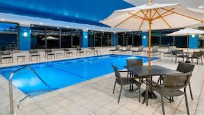 An indoor swimming pool area with tiled flooring, lounge chairs, and tables with umbrellas.