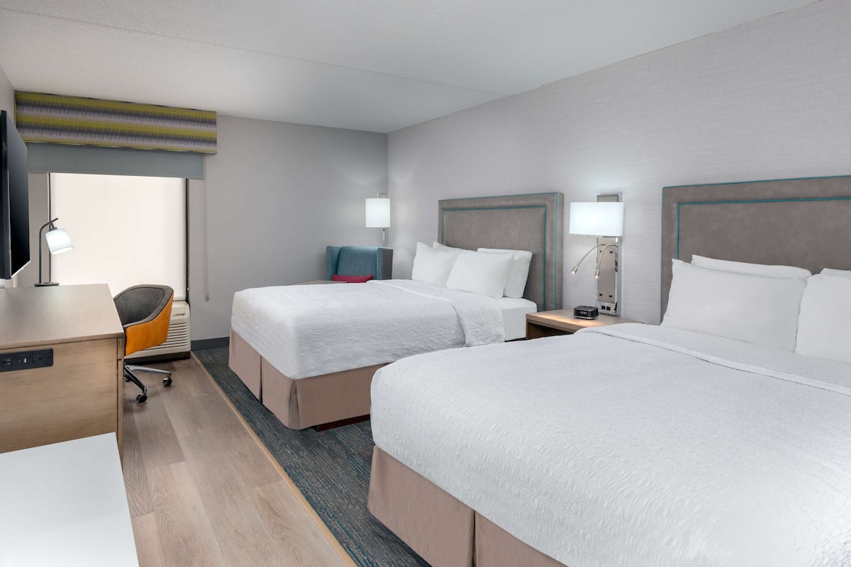 A hotel room with two beds, a desk, a chair, and a TV.