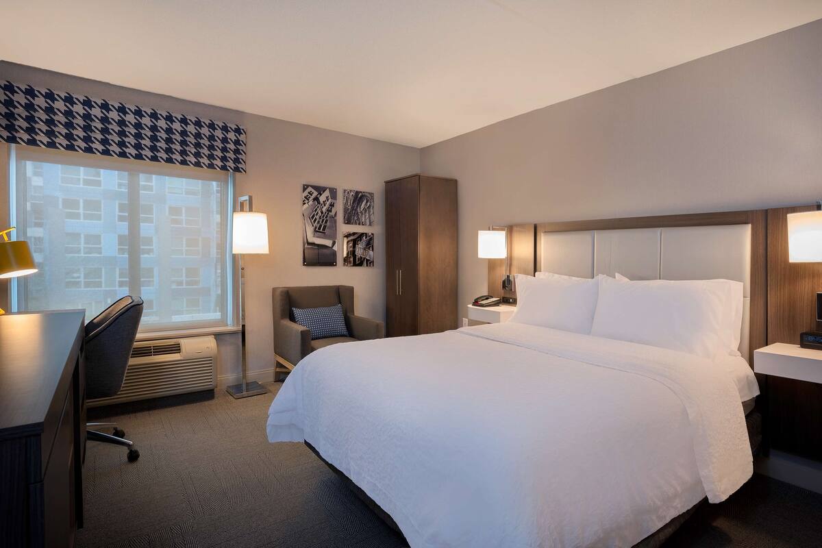 A hotel room with a large bed, two bedside tables with lamps, a desk with a chair, a radiator, and a window with a view of buildings.
